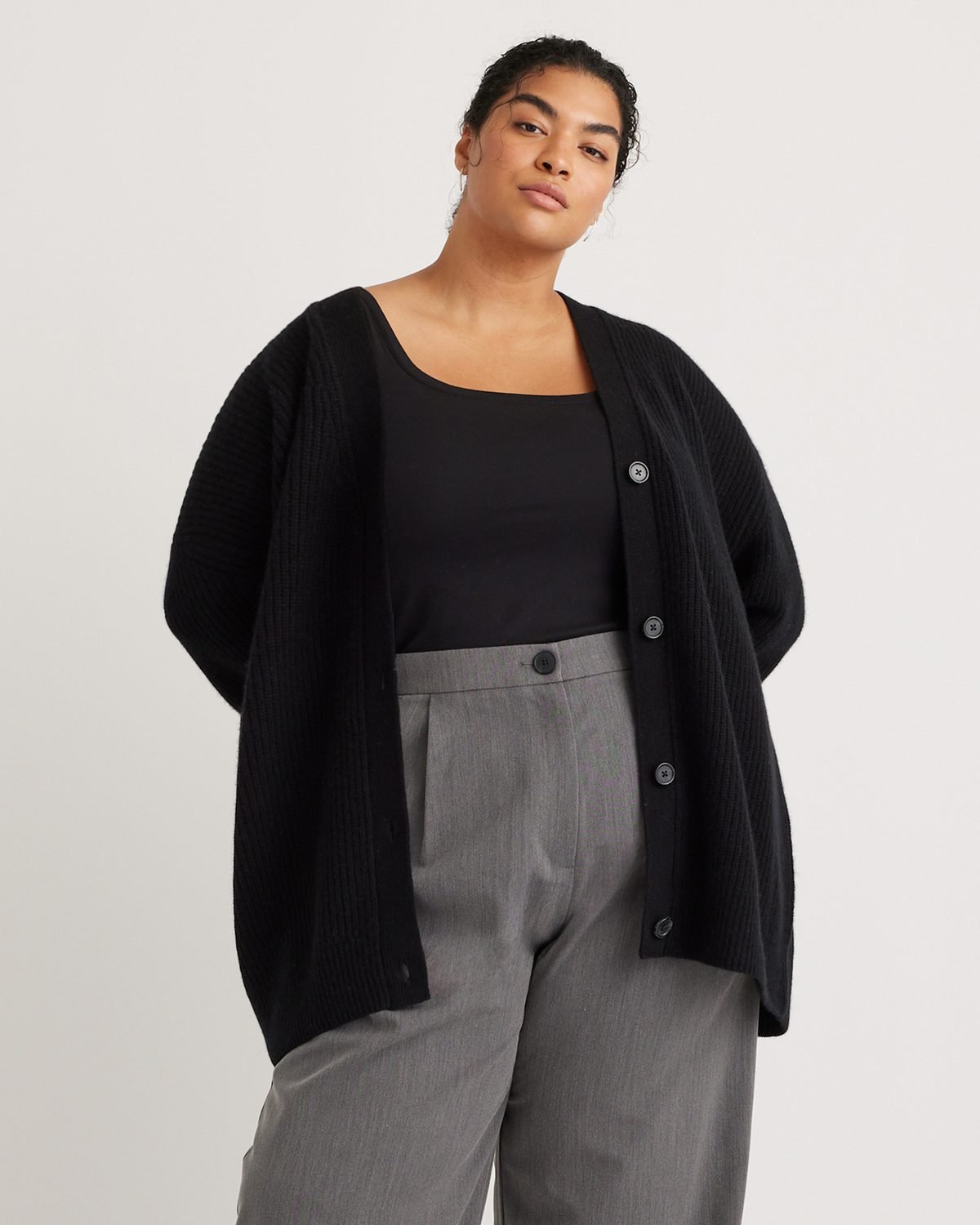 Chunky black cashmere boyfriend cardigan worn open over a tank with grey trousers, worn by a woman over 50.