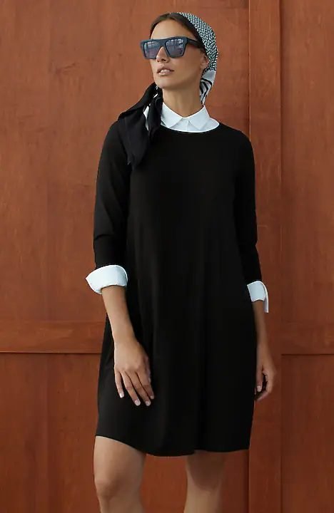 Black knit A line dress with three quarter sleeves and back pleats, shown layered over a white collared shirt.